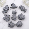 A charcoal grey collection of eight handmade vegan Halloween soaps featuring a bat, witch's hat, star-patterned sweet, haunted house, black cat, jack-o'-lantern, and a ghost, arranged on a light wooden surface with white floral accents.