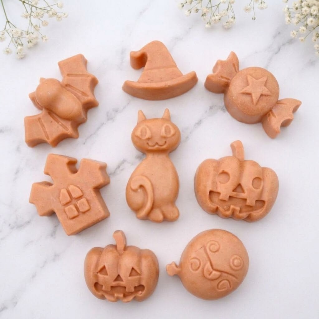 A terracotta collection of eight handmade vegan Halloween soaps featuring a bat, witch's hat, star-patterned sweet, haunted house, black cat, jack-o'-lantern, and a ghost, arranged on a light wooden surface with white floral accents.