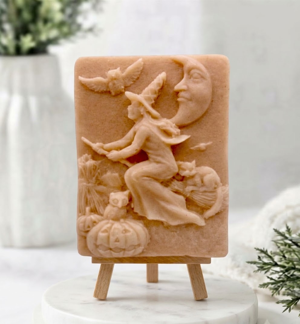 A terracotta handmade vegan soap bar featuring a detailed relief carving of a witch flying on a broomstick past a crescent moon, owls, and pumpkins.