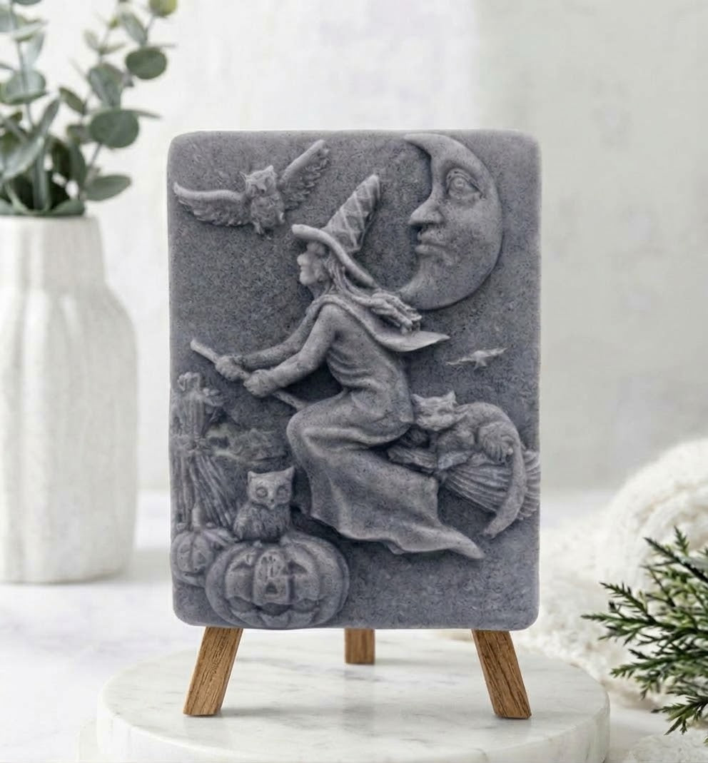 A charcoal grey handmade vegan soap bar featuring a detailed relief carving of a witch flying on a broomstick past a crescent moon, owls, and pumpkins.