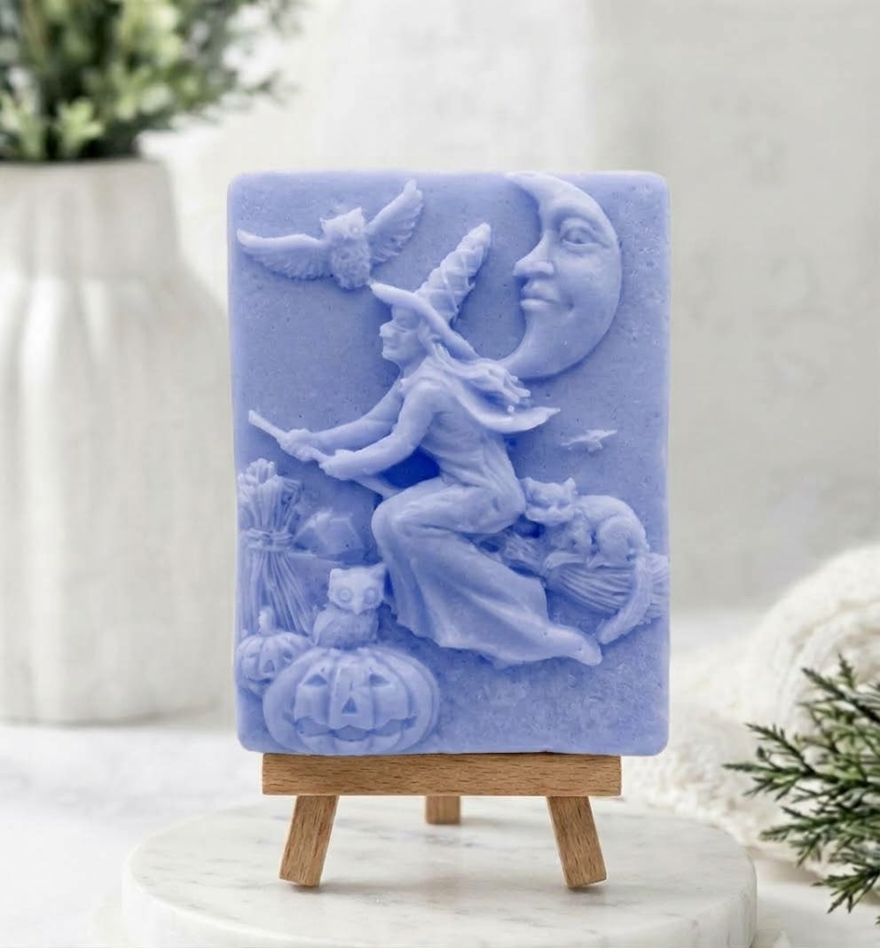 A soft blue handmade vegan soap bar featuring a detailed relief carving of a witch flying on a broomstick past a crescent moon, owls, and pumpkins.