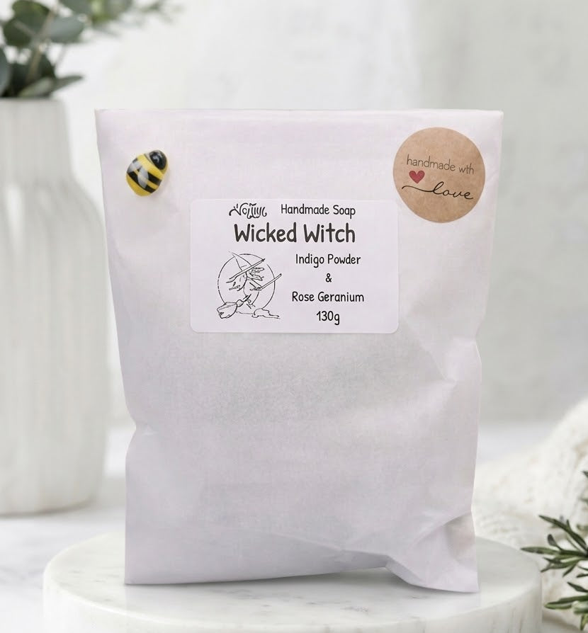 A white paper pouch of 'Wicked Witch' handmade soap by 'Magic Shop' sits on a white marble coaster. The background is a soft, bright, and slightly out-of-focus setting with a white vase of greenery on the left and a white knit blanket on the right.