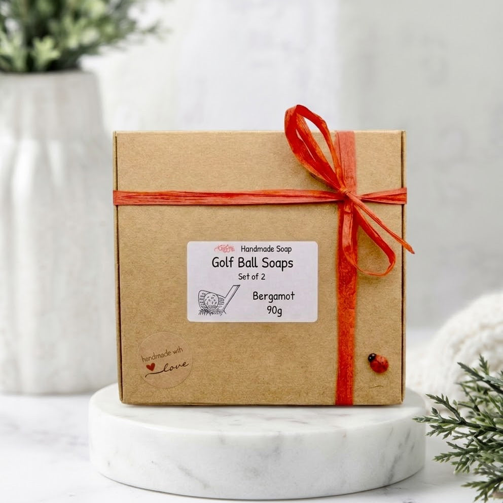 A square kraft paper box of Handmade Golf Ball Soaps, Set of 2, Bergamot, tied with a red ribbon and bow, displayed on a white marble pedestal.