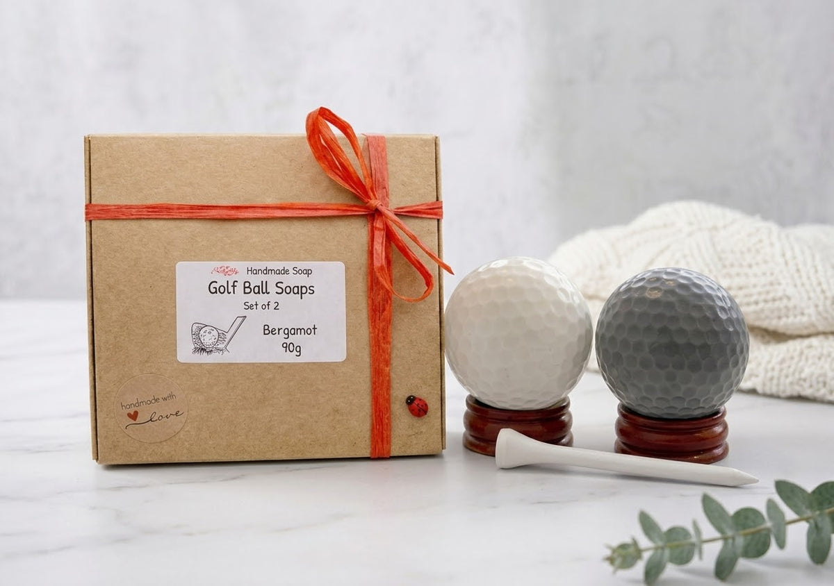A gift set of two Bergamot golf ball soaps in their kraft paper packaging box with a red ribbon, displayed next to the actual soaps shaped like white and gray golf balls on wooden stands with a white tees accessory on a marble surface.