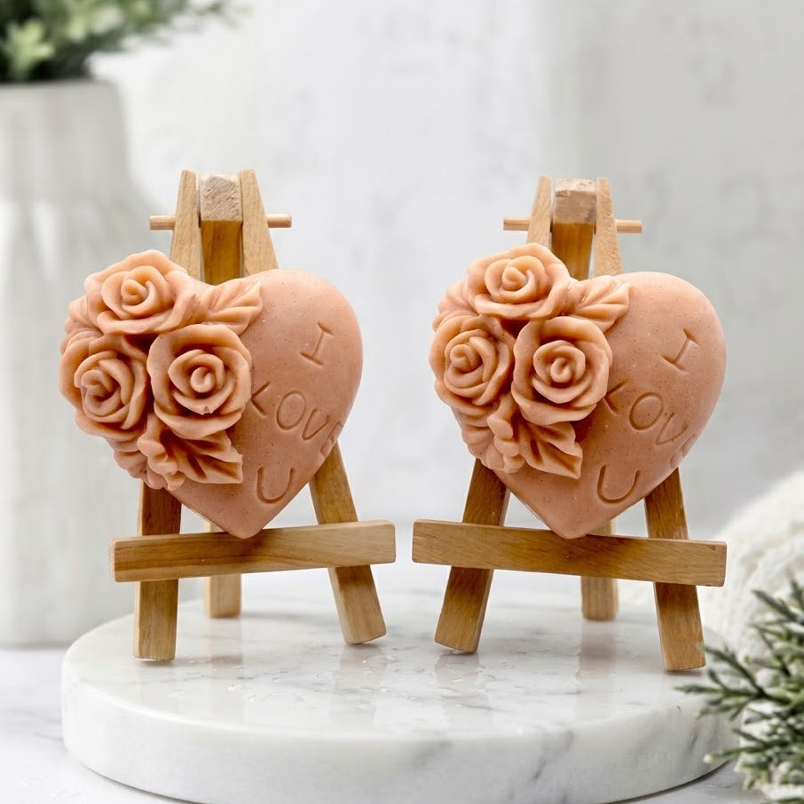 Two terracotta heart-shaped decorative soap bars engraved with 'I LOVE YOU' and 'I LOVE U', each adorned with raised rose details, displayed on a small wooden easel and beside one standing upright on a light surface with soft floral props.