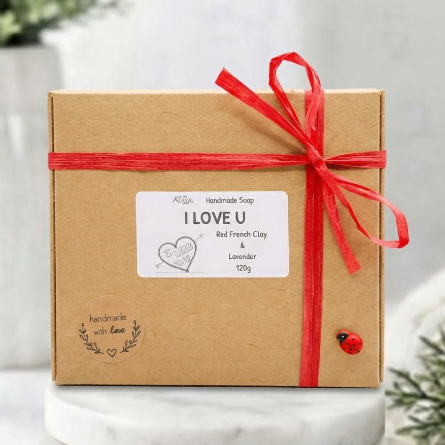 A square kraft paper gift box tied with a red ribbon and bow, featuring a white label that reads 'I LOVE U' and 'Handmade Soap Red French Clay & Lavender 120g'. The box is photographed on a white marble pedestal with soft-focus greenery in the background.