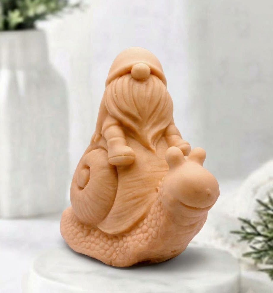 A terracotta handmade vegan soap gift, sculpted as a whimsical gnome (gonk) with a tall, slouchy hat and a long beard, sitting on the shell of a large snail with a textured, bumpy body.