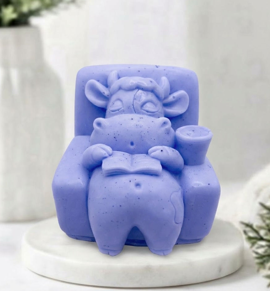 A lavender blue soap bar shaped like a sleeping cow sitting in an armchair reading a book, displayed on a white marble round tray with a blurred background.