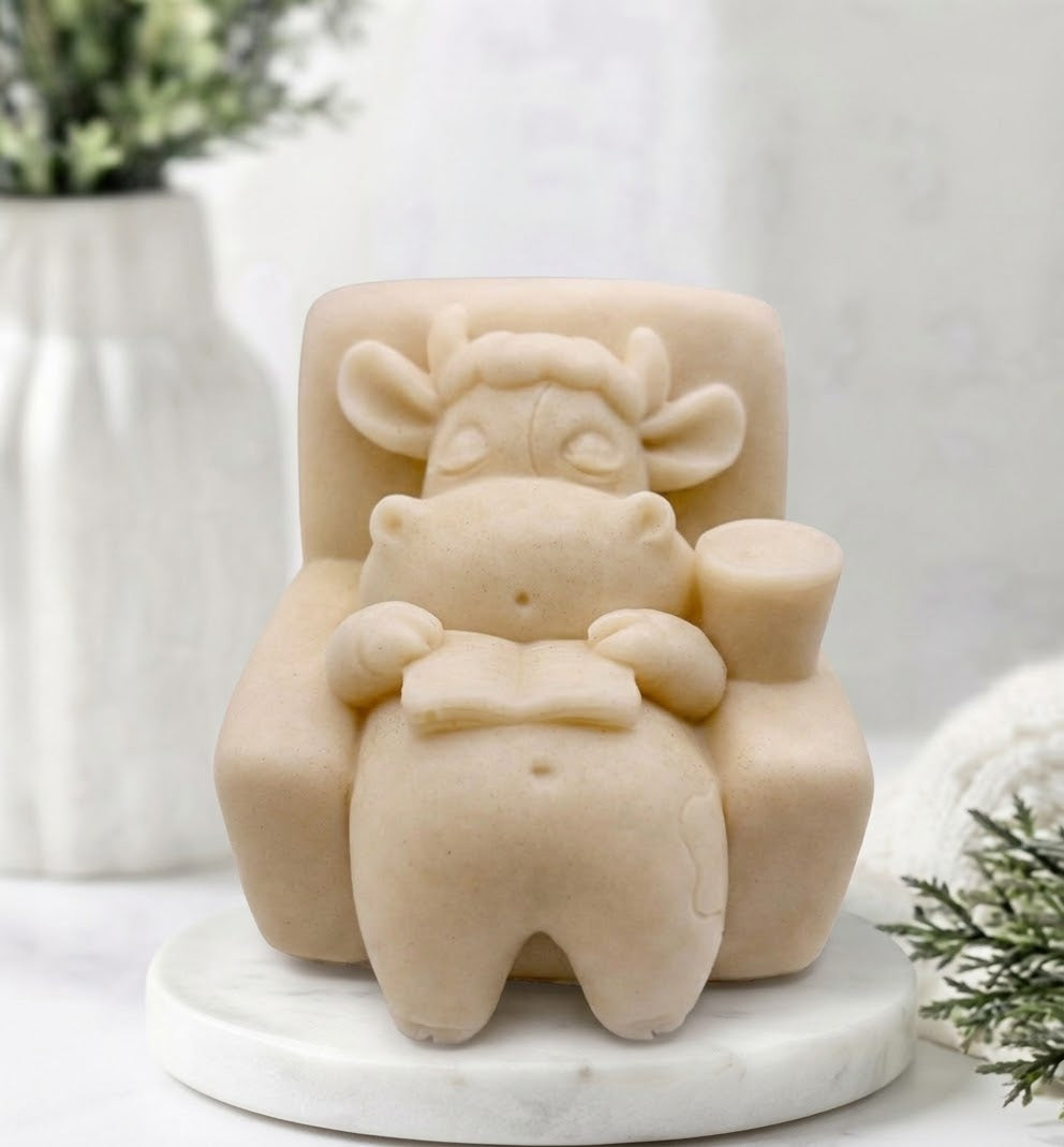 A soft yellow sculpted soap of a cow sitting in a chair and reading a book, displayed on a white marble base with a soft, blurred background.
