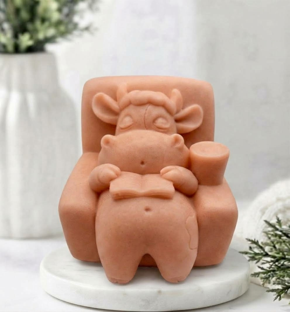 A terracotta coloured novelty soap figurine of a lazy cow sitting in an armchair and reading a book, displayed on a white marble base with a soft-focus background.