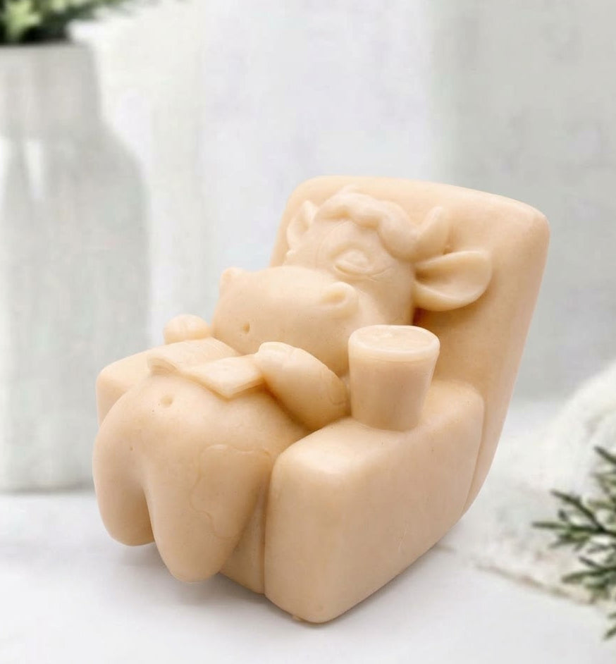 A soft yellow novelty soap bar shaped like a cow lounging in a chair, displayed on a white surface with a blurred white vase and greenery in the background.