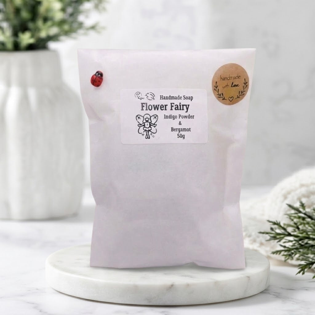 A paper bag of 'Flower Fairy' handmade soap sits on a white marble coaster. The bag is pale pink with a white label and a round kraft paper sticker.