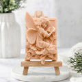 A soft pink soap bar sculpted with a fairy and flowers, displayed on a small wooden easel stand over a white marble base.