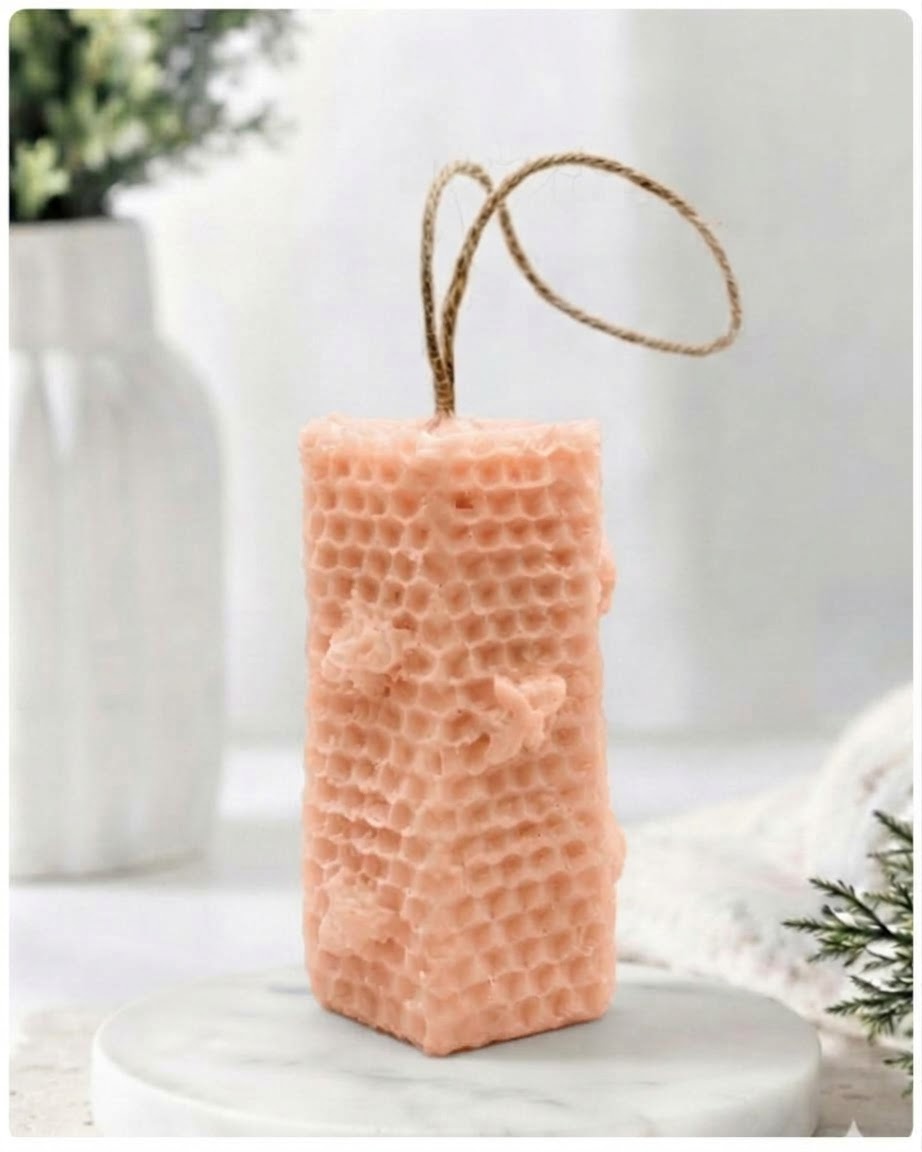 A pink-coloured honeycomb vegan soap with a natural jute hanging loop, displayed on a white marble pedestal against a soft, bright, blurred background.