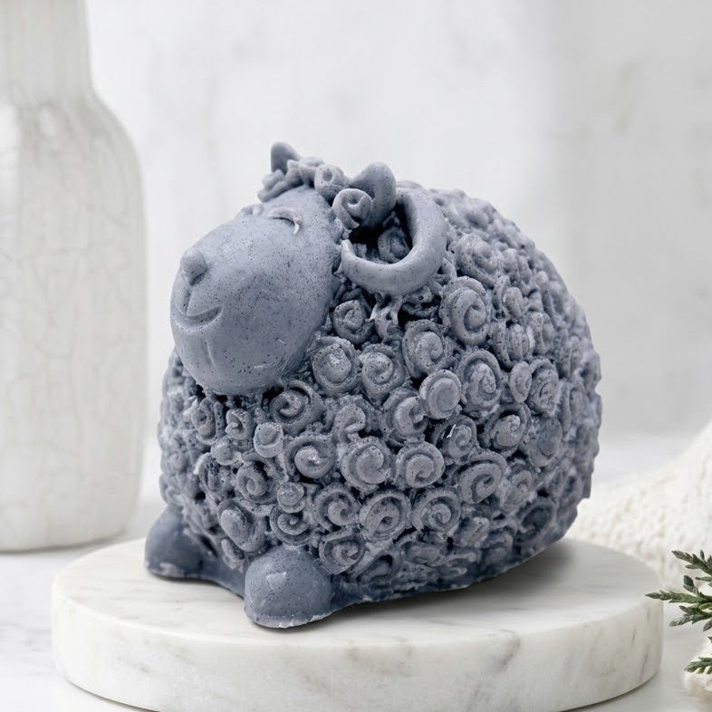 A grey textured sheep soap with curly wool sits on a round white marble tray, styled on a marble surface with a white ceramic vase and greenery in the background.