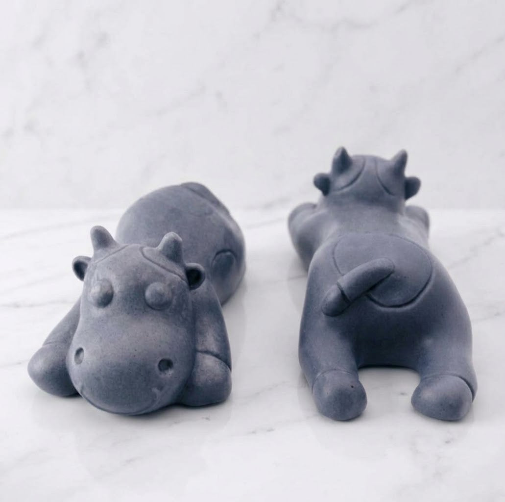 Two charcoal grey sleeping cow shaped soaps are resting side-by-side on a white marble countertop.