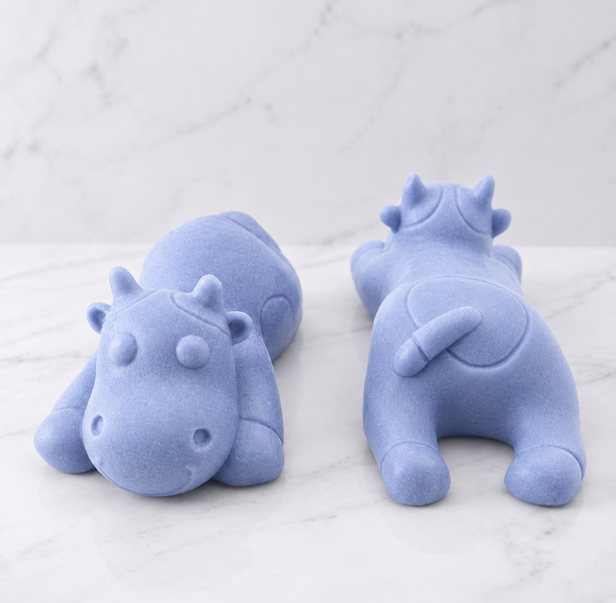 Two soft blue sleeping cow shaped soaps are resting side-by-side on a white marble countertop.