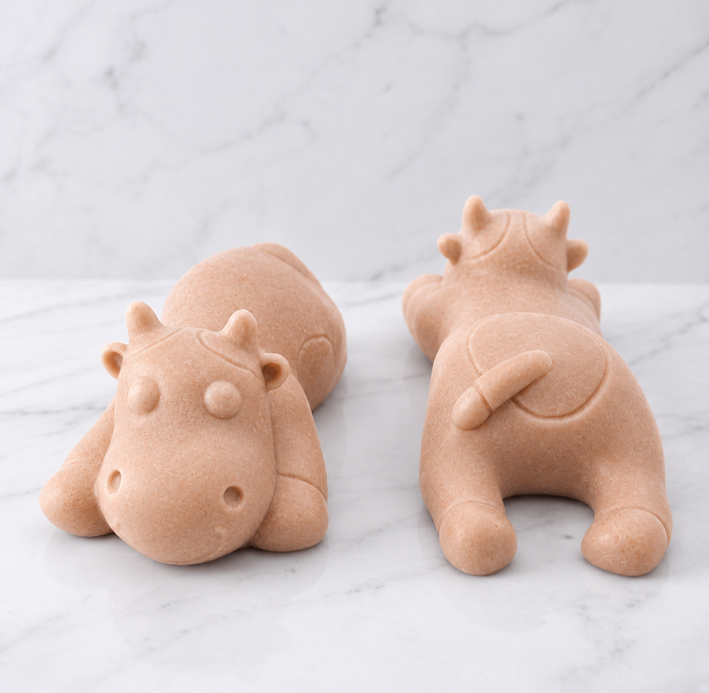 Two soft pink sleeping cow shaped soaps are resting side-by-side on a white marble countertop.