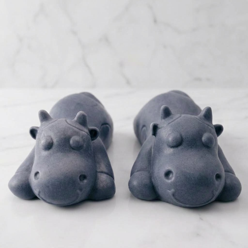 Two forward facing charcoal grey sleeping cow shaped soaps are resting side-by-side on a white marble countertop.