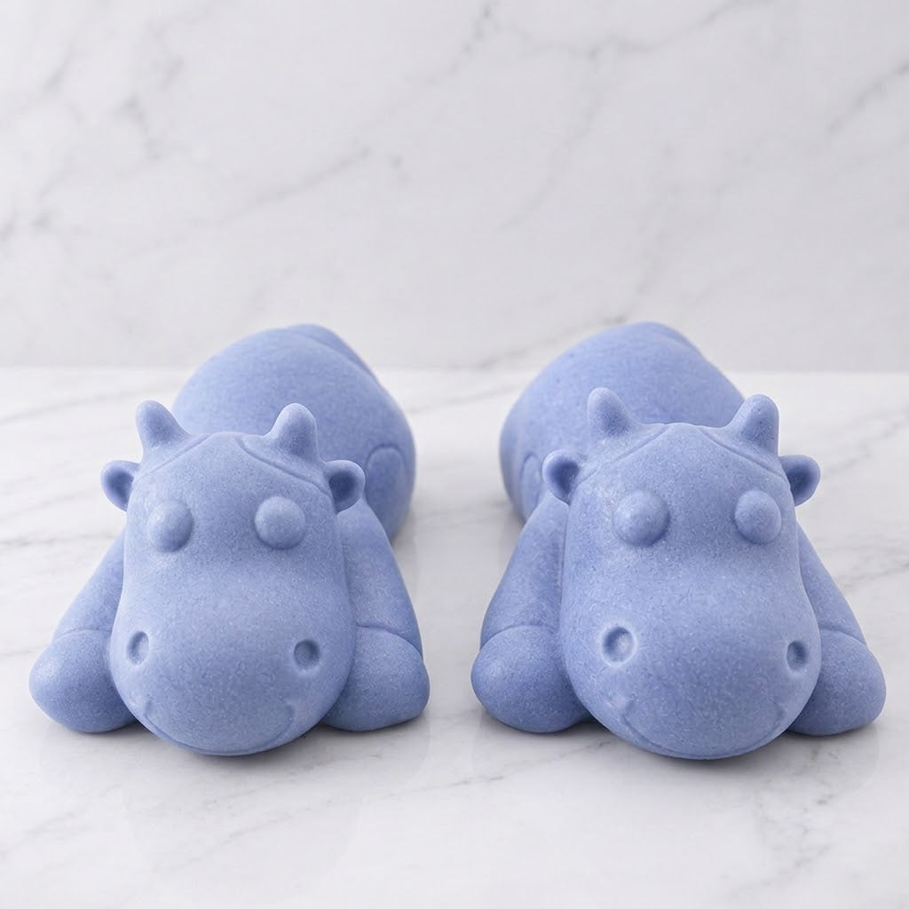 Two forward facing soft blue sleeping cow shaped soaps are resting side-by-side on a white marble countertop.