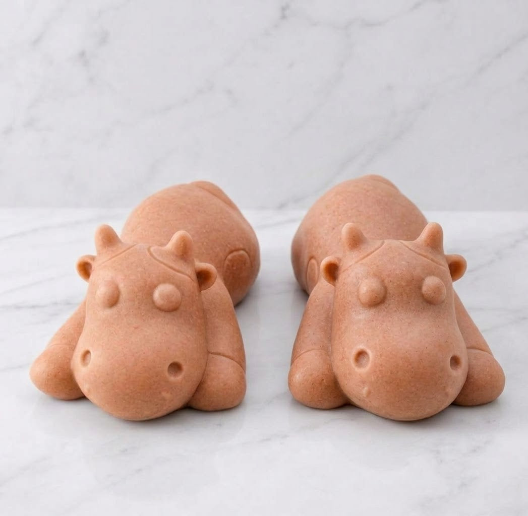 Two forward facing terracotta coloured sleeping cow shaped soaps are resting side-by-side on a white marble countertop.