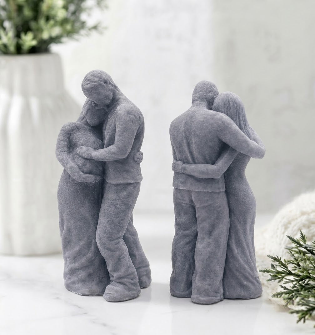 A front and rear view of a handmade soap sculpture. It depicts a man in pants and a shirt embracing a pregnant woman in a long dress from behind, with his hands over her belly. They are on a round marble stand