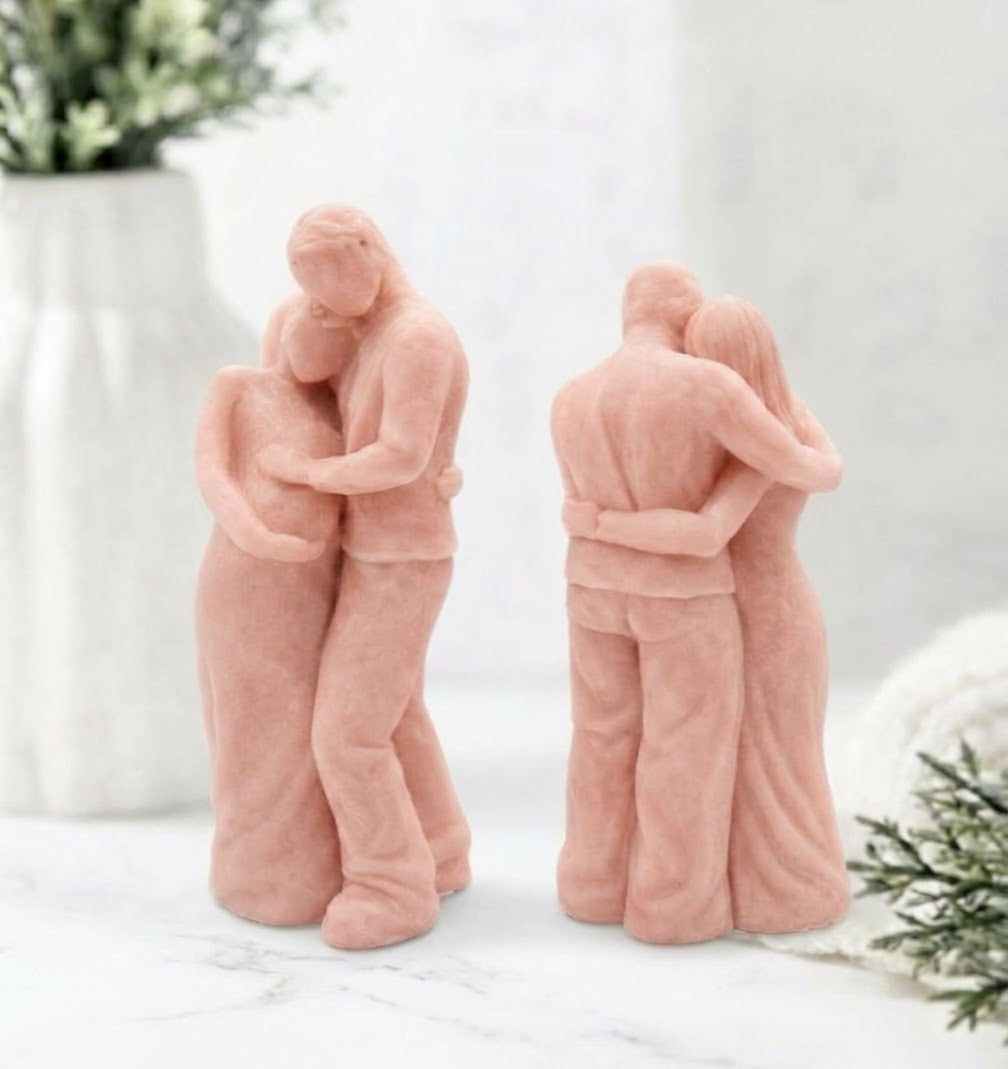 A front and back view of a soft pink soap sculpture. It depicts a man in pants and a shirt embracing a pregnant woman in a long dress from behind, with his hands over her belly. They are on a round marble stand