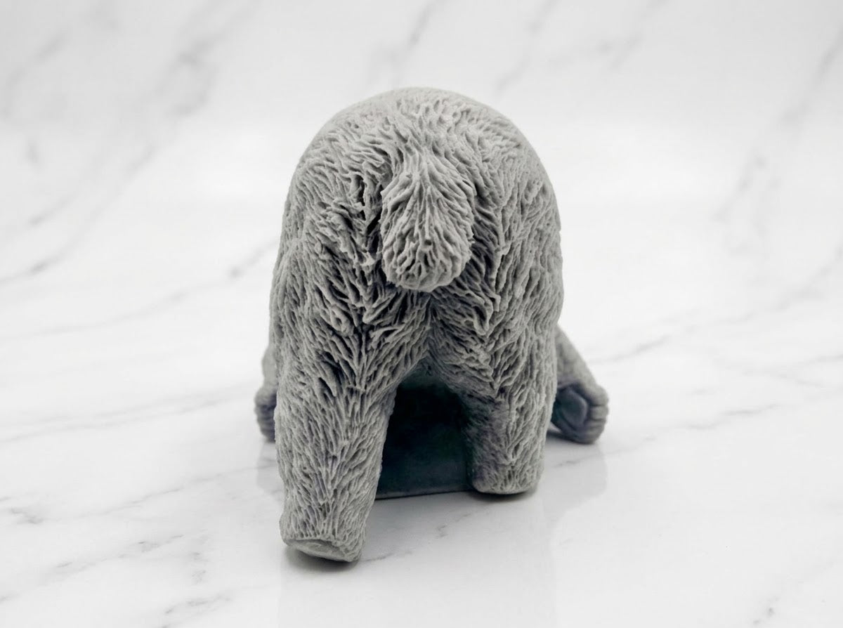 A rear view of a charcoal-grey bar of soap sculpted into the form of a sleeping polar bear lying on its side, resting on white marble.
