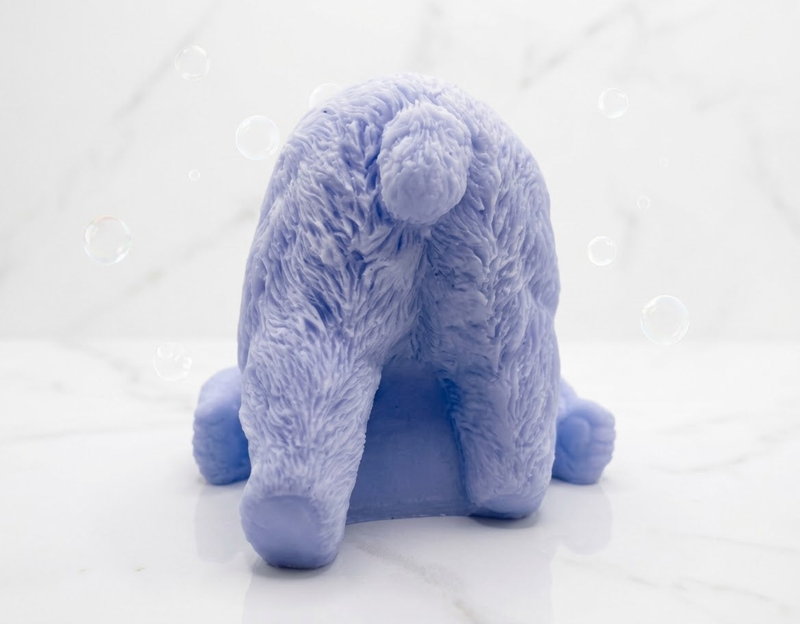 A rear view of a soft blue bar of soap sculpted into a lazy polar bear figure lying down on its side, set against a white marble background.