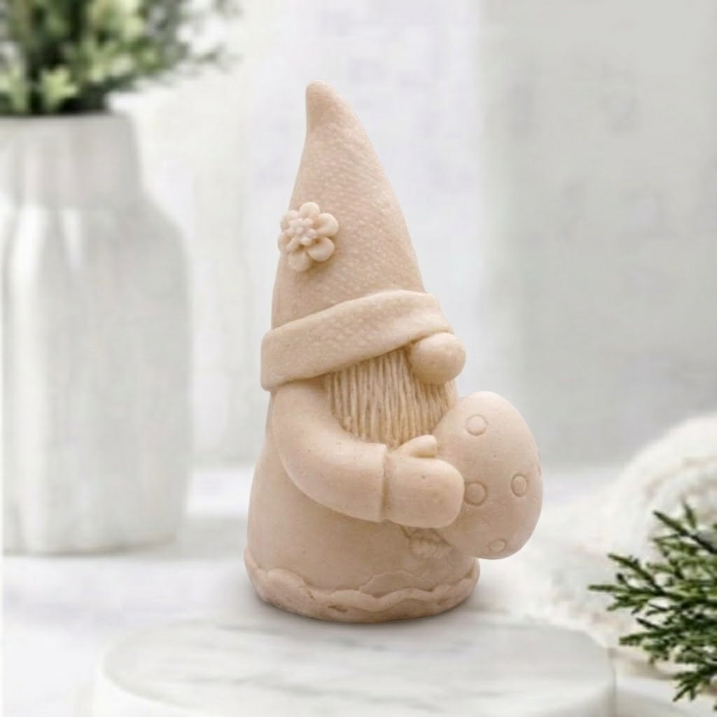 A side view of a soft yellow detailed handmade vegan soap gift,  sculpted as a whimsical Easter gnome (gonk) wearing a tall pointed hat with a flower detail, holding a large patterned Easter egg.
