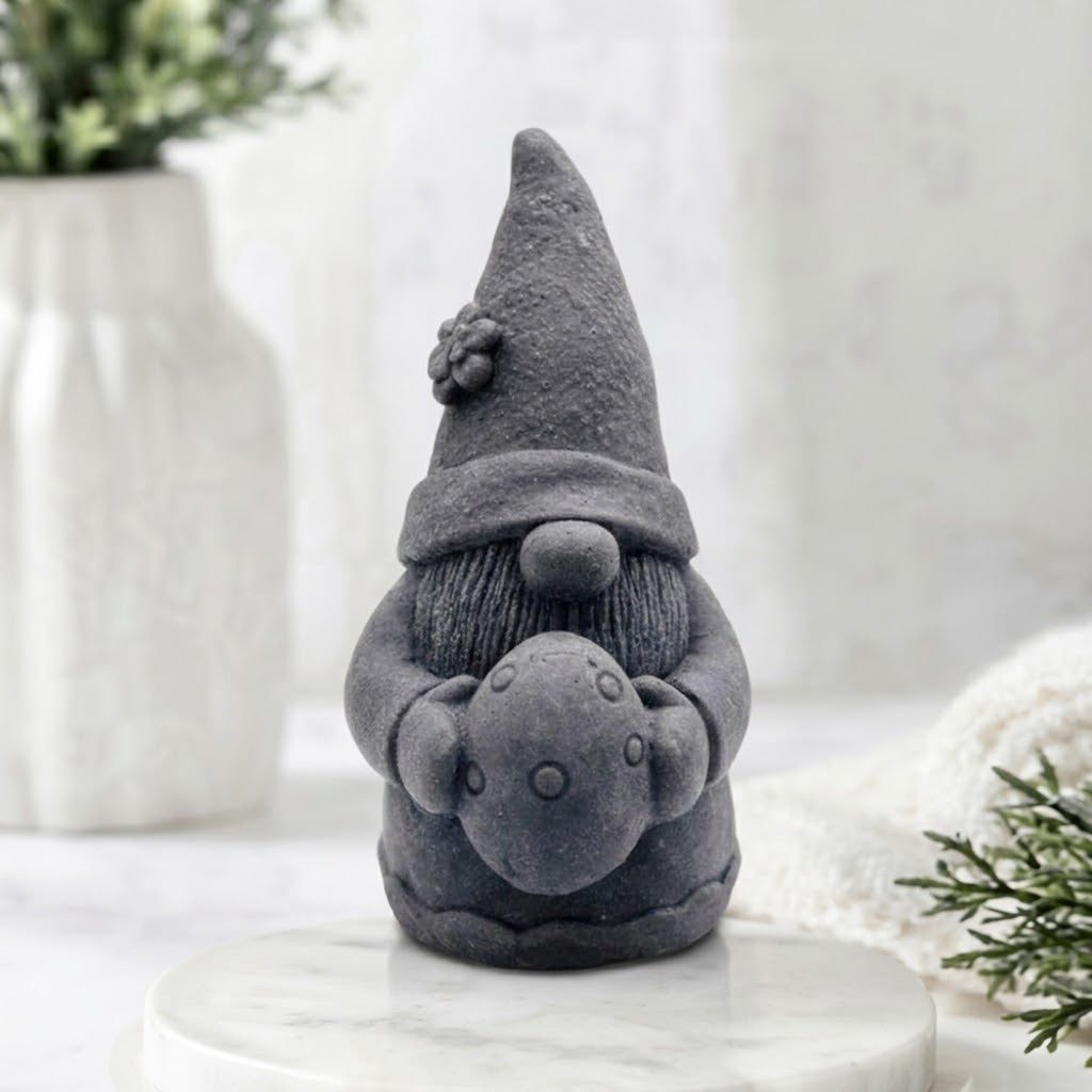 A charcoal grey detailed handmade vegan soap gift,  sculpted as a whimsical Easter gnome (gonk) wearing a tall pointed hat with a flower detail, holding a large patterned Easter egg.