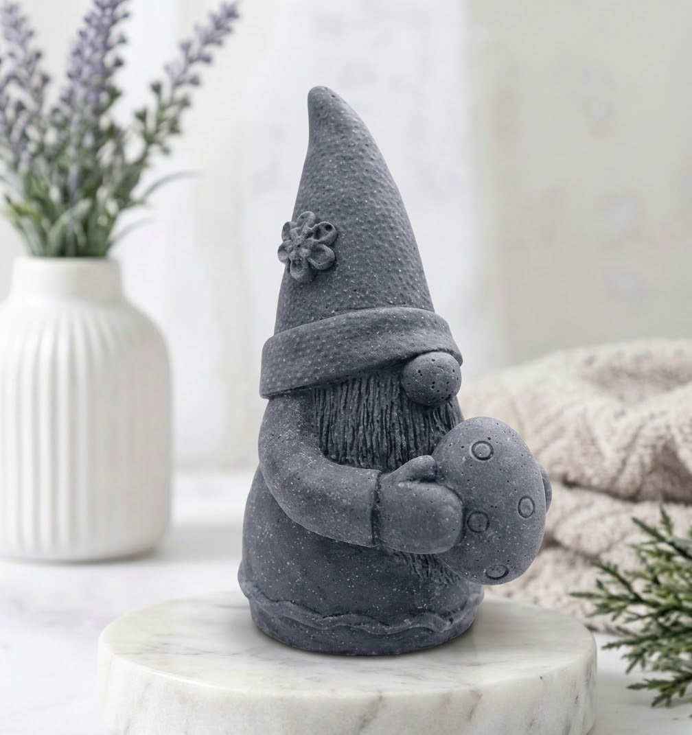 A charcoal grey gnome soap holding a small egg, displayed on a round white marble pedestal with a white ribbed vase of lavender in the background 