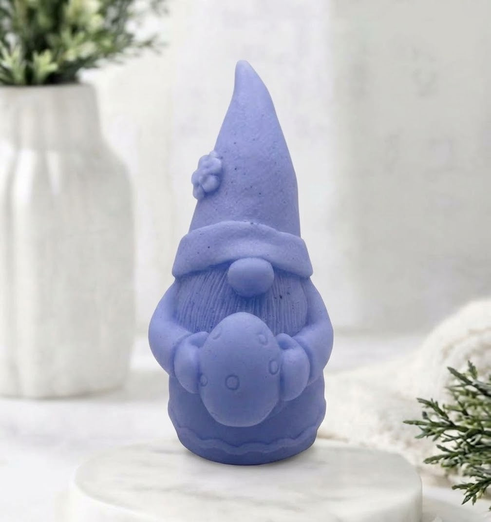 A detailed lavender blue coloured handmade vegan soap gift,  sculpted as a whimsical Easter gnome (gonk) wearing a tall pointed hat with a flower detail, holding a large patterned Easter egg.