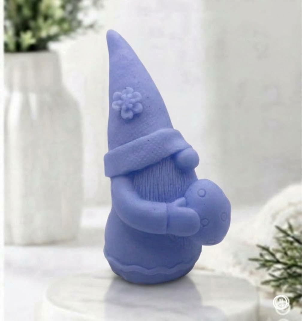 A side view of a soft blue detailed handmade vegan soap gift, sculpted as a whimsical Easter gnome (gonk) wearing a tall pointed hat with a flower detail, holding a large patterned Easter egg and a flower in his hat. 