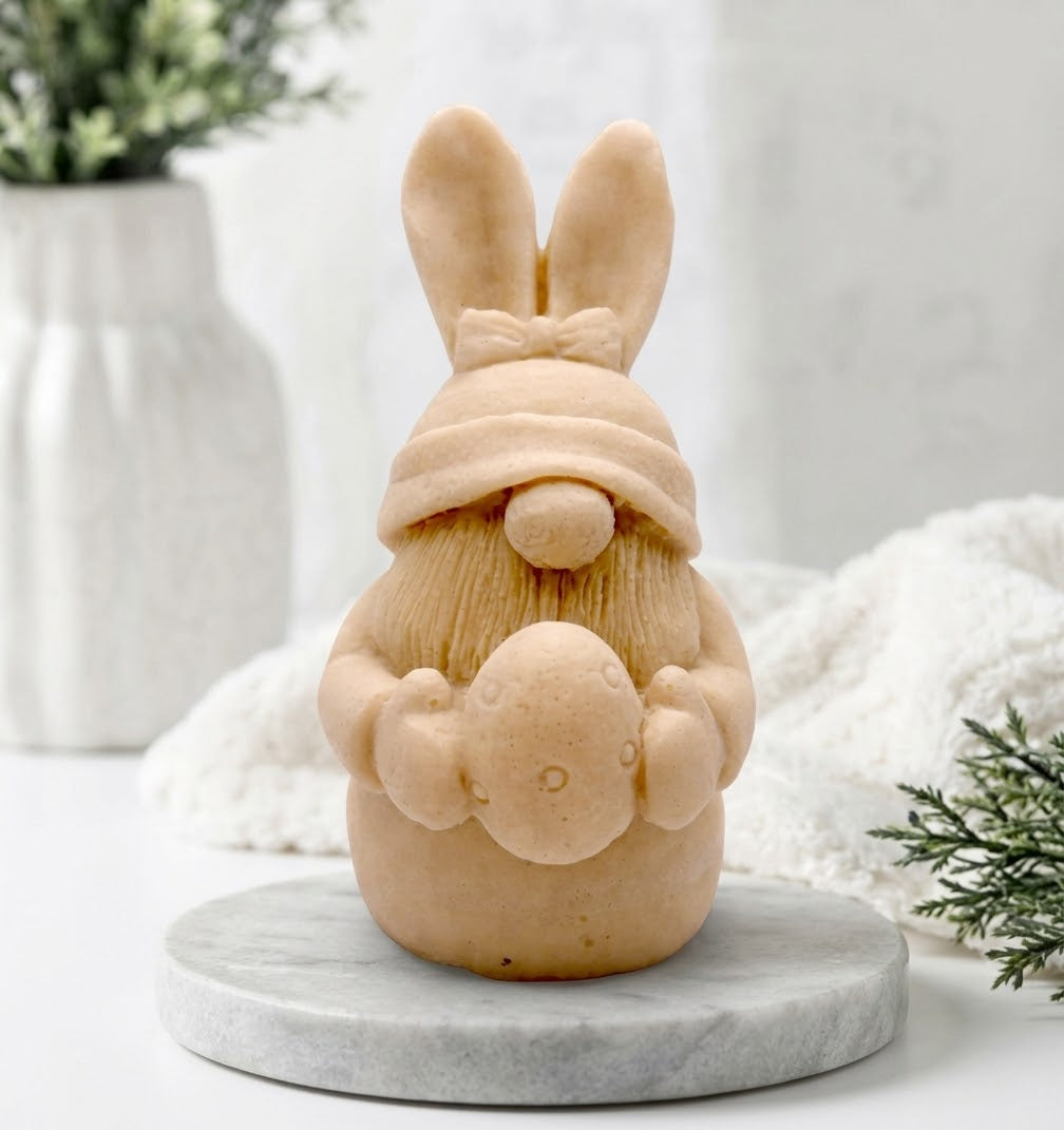 A soft yellow coloured detailed handmade vegan soap in a natural colour, sculpted as a whimsical Easter gnome (gonk) with long rabbit ears and a bow on its hat, holding a patterned Easter egg, displayed on a light wooden surface. Easter gift 