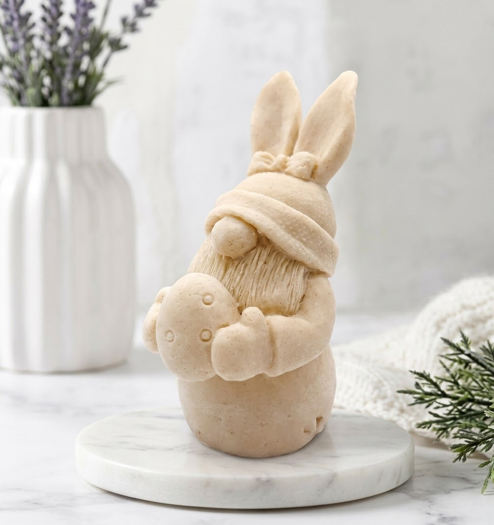 A soft yellow Easter bunny gnome soap wearing a knit hat and holding a bunny, displayed on a round white marble coaster with a white vase of lavender and greenery in the background.