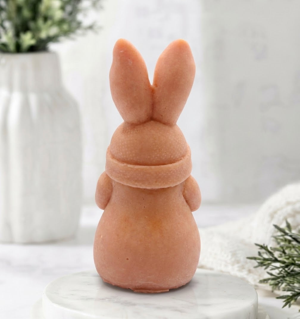 A rear view of a detailed handmade vegan soap in a natural colour, sculpted as a whimsical Easter gnome (gonk) with long rabbit ears and a bow on its hat, holding a patterned Easter egg, displayed on a light wooden surface. Easter gift 