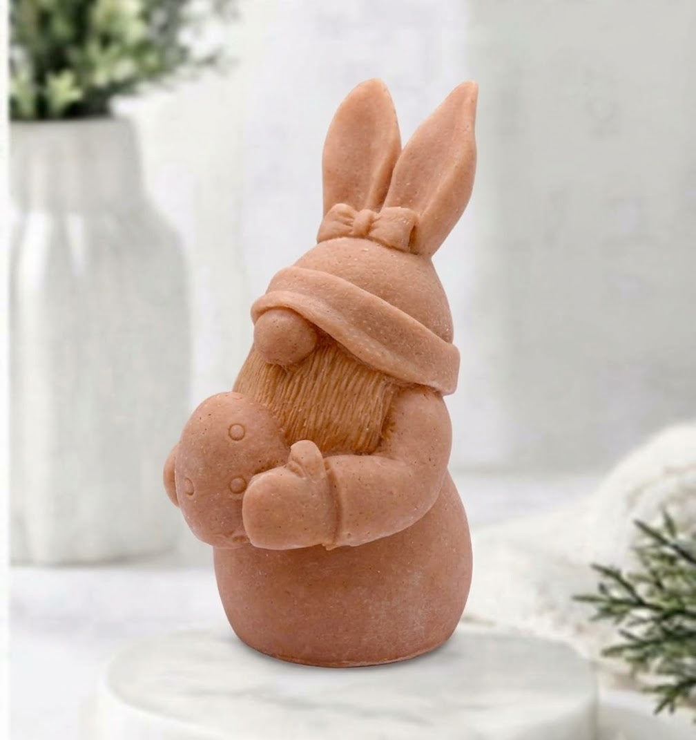 A terracotta-coloured Easter bunny gnome soap wearing a knit hat and holding a small egg, displayed on a white marble pedestal with a soft-focus background.