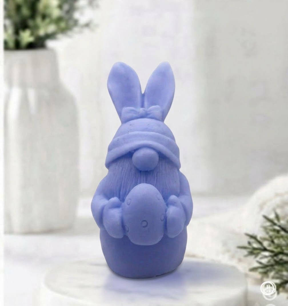 A lavender blue Easter bunny soap wearing a knit hat and holding an egg, displayed on a white pedestal with a blurred background of a vase and greenery.