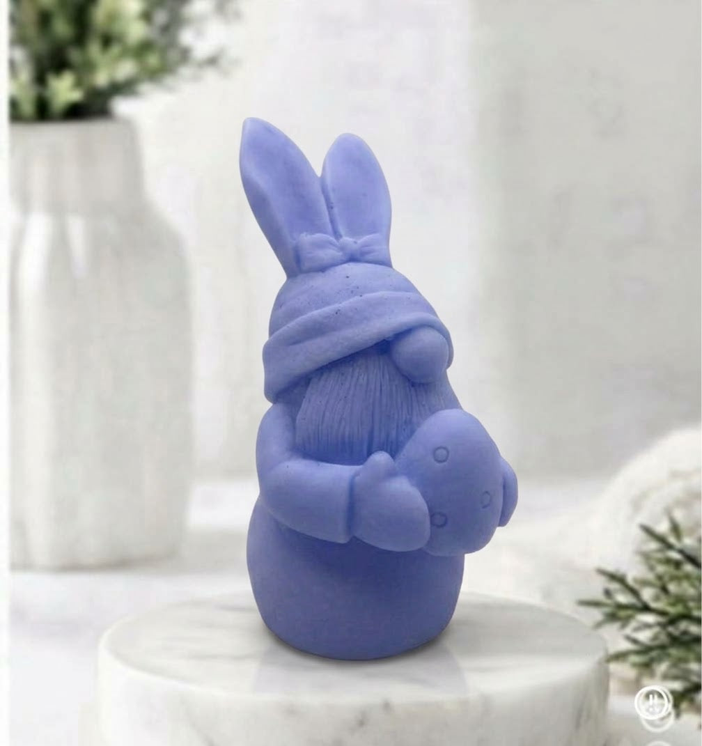 A side view of a blue detailed handmade vegan soap in a natural colour, sculpted as a whimsical Easter gnome (gonk) with long rabbit ears and a bow on its hat, holding a patterned Easter egg, displayed on a light wooden surface. Easter gift 