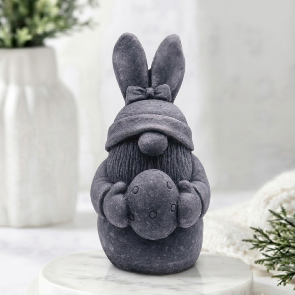 A charcoal grey Easter rabbit gnome soap wearing a knit hat and holding an egg, displayed on a white marble pedestal with a softly blurred home interior background.