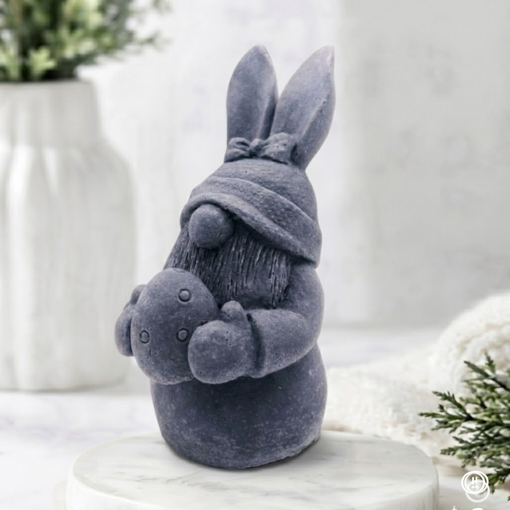 A side view of a grey detailed handmade vegan soap in a natural colour, sculpted as a whimsical Easter gnome (gonk) with long rabbit ears and a bow on its hat, holding a patterned Easter egg, displayed on a light wooden surface. Easter gift 