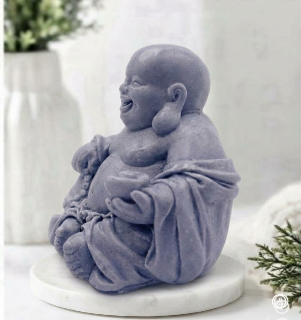 A detailed, handcrafted charcoal grey soap bar sculpted into the form of a seated, smiling Laughing Buddha. The Buddha is cross-legged on a white marble disc and holds a string of prayer beads in its left hand and an offering bowl in its right, set against a detailed spa bathroom background.
