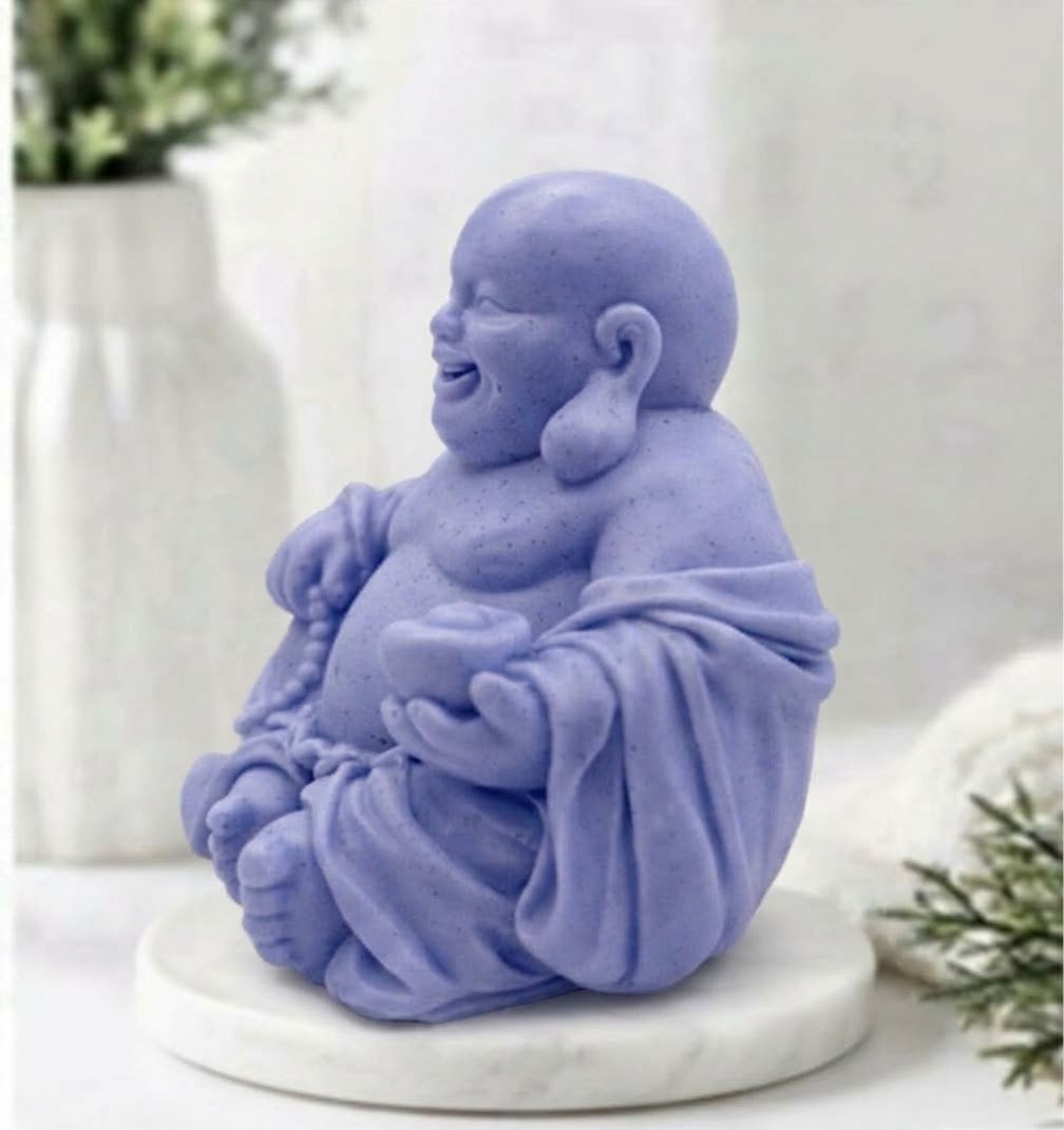 A detailed, handcrafted blue coloured soap bar sculpted into the form of a seated, smiling Laughing Buddha. The Buddha is cross-legged on a white marble disc and holds a string of prayer beads in its left hand and an offering bowl in its right, set against a detailed spa bathroom background