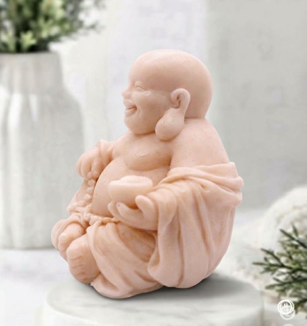 A detailed, handcrafted pink coloured soap bar sculpted into the form of a seated, smiling Laughing Buddha. The Buddha is cross-legged on a white marble disc and holds a string of prayer beads in its left hand and an offering bowl in its right, set against a detailed spa bathroom background 