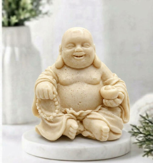 A detailed, handcrafted soft yellow coloured soap bar, sculpted into the form of a seated, smiling Laughing Buddha. The Buddha is cross-legged on a white marble disc and holds a string of prayer beads in its left hand and an offering bowl in its right, set against a detailed spa bathroom background