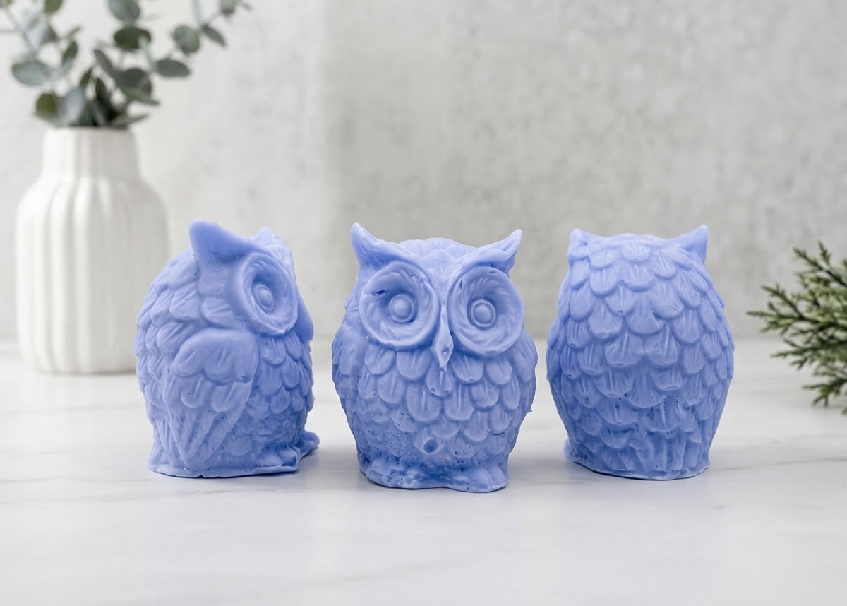 A gift set featuring three soft blue handmade vegan soaps shaped like barn owls , arranged in a row on a light wooden surface with white flowers in the background 