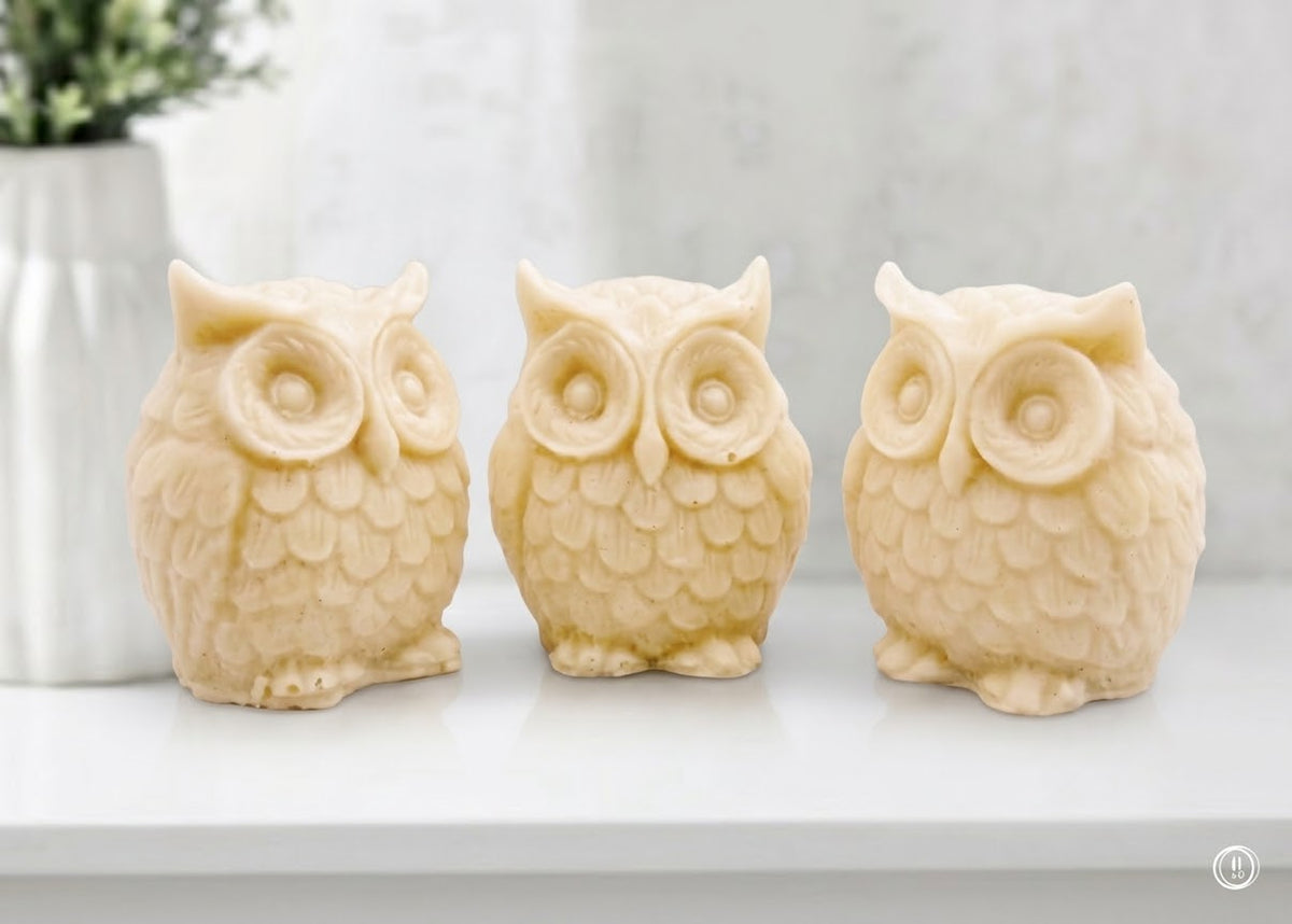 A gift set featuring three soft yellow handmade vegan soaps shaped like barn owls , arranged in a row on a light wooden surface with white flowers in the background 