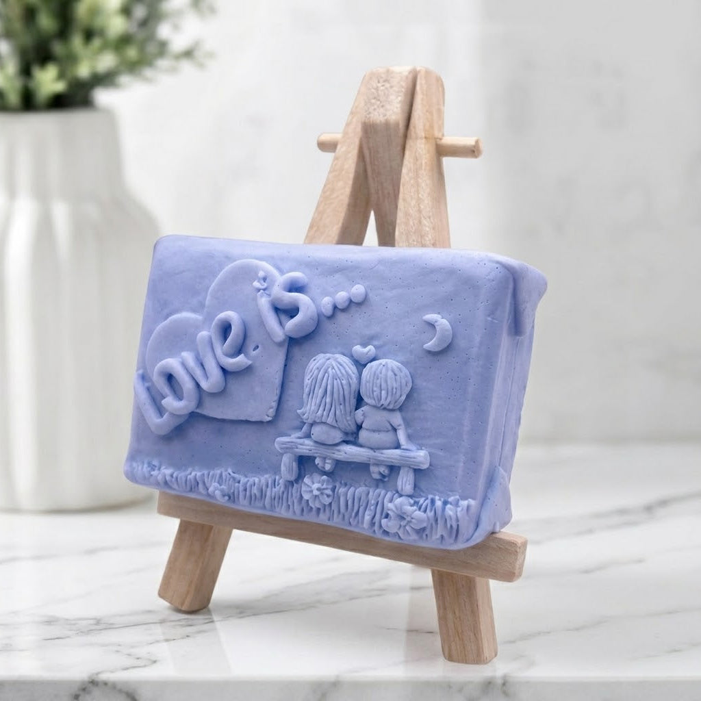 A rectangular handmade soft blue vegan soap featuring a detailed 3D relief carving of a young couple sitting on a bench under a crescent moon, with the words "Love is..." embossed inside a heart, displayed on a wooden easel.