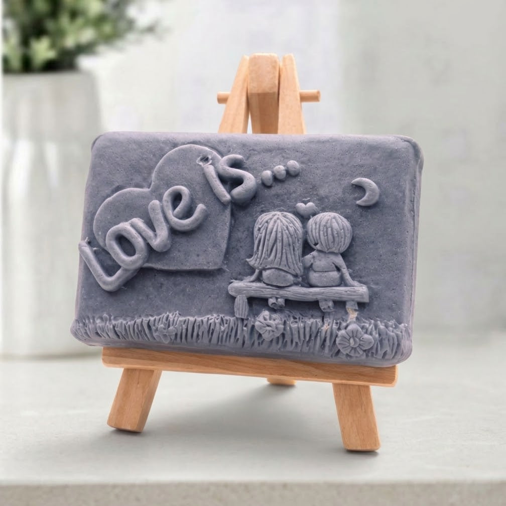 A rectangular charcoal handmade vegan soap featuring a detailed 3D relief carving of a young couple sitting on a bench under a crescent moon, with the words "Love is..." embossed inside a heart, displayed on a wooden easel.